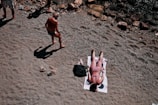 A person is sunbathing on a towel at a pebbly beach. Another person is walking nearby, casting a long shadow on the ground. Surrounding them are rocks and sand, creating a natural setting.