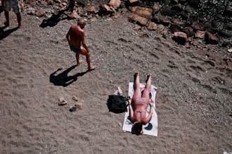 A person is sunbathing on a towel at a pebbly beach. Another person is walking nearby, casting a long shadow on the ground. Surrounding them are rocks and sand, creating a natural setting.