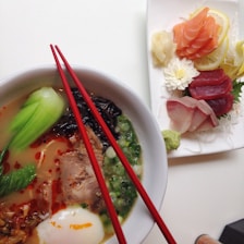 A bowl of ramen with rich broth, topped with slices of meat, green vegetables, an egg, seaweed, and green onions, accompanied by red chopsticks resting across the bowl. Beside it, a plate of assorted sashimi including salmon, tuna, and other types of fish neatly arranged with lemon slices, flowers, and wasabi.