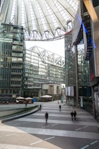 A modern architectural space with a large, airy atrium and a distinctive, conical-shaped roof structure. Surrounding glass buildings reflect natural light into the open plaza below, where a few people are walking. Cafés with outdoor seating are arranged around the edges, adding a casual atmosphere.