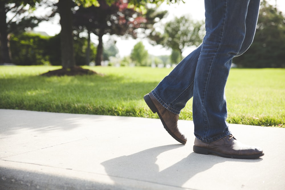 갈색 신발과 파란색 데님 청바지를 입은 사람이 낮에 푸른 잔디밭 근처의 콘크리트 땅을 걷고 있습니다
