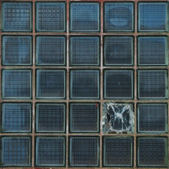 A grid of square glass blocks, one of which is cracked and broken. The glass blocks have a blue tint and are arranged in a five-by-five formation. The cracked block has visible lines and patterns of the shattered glass in contrast to the rest of the uniform blocks.