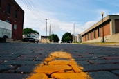 A low-angle view of a brick-paved street with a yellow line running through the center. On the left, a white service vehicle is parked next to a red brick building. On the right, a modern building with a bike parked in front is visible. Utility poles and wires stretch across the sky, and trees and distant structures can be seen further down the street.