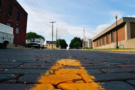 A low-angle view of a brick-paved street with a yellow line running through the center. On the left, a white service vehicle is parked next to a red brick building. On the right, a modern building with a bike parked in front is visible. Utility poles and wires stretch across the sky, and trees and distant structures can be seen further down the street.