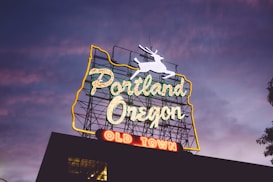 A neon sign prominently displaying 'Portland Oregon' with 'Old Town' in red below. The sign is outlined with a map-like shape and features a white stag leaping at the top. It is set against a twilight sky with shades of purple and blue.