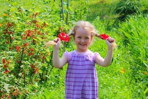 A young girl with braided hair stands in a lush garden, holding red flowers in her hands. She is smiling broadly and wears a purple, striped dress. The background is filled with vibrant green foliage and clusters of red berries.
