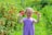 A young girl with braided hair stands in a lush garden, holding red flowers in her hands. She is smiling broadly and wears a purple, striped dress. The background is filled with vibrant green foliage and clusters of red berries.
