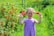A young girl with braided hair stands in a lush garden, holding red flowers in her hands. She is smiling broadly and wears a purple, striped dress. The background is filled with vibrant green foliage and clusters of red berries.