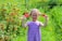 A young girl with braided hair stands in a lush garden, holding red flowers in her hands. She is smiling broadly and wears a purple, striped dress. The background is filled with vibrant green foliage and clusters of red berries.