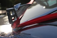 A shiny metal car emblem with wings is adorned with a red ribbon. The emblem reflects sunlight, creating bright, gleaming highlights on the surface of the car hood.