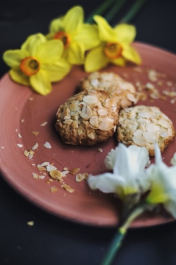 Three almond-topped cookies are placed on a pink plate, surrounded by yellow daffodil flowers. Almond flakes are scattered around the cookies, adding a rustic charm.