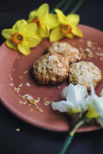 Three almond-topped cookies are placed on a pink plate, surrounded by yellow daffodil flowers. Almond flakes are scattered around the cookies, adding a rustic charm.