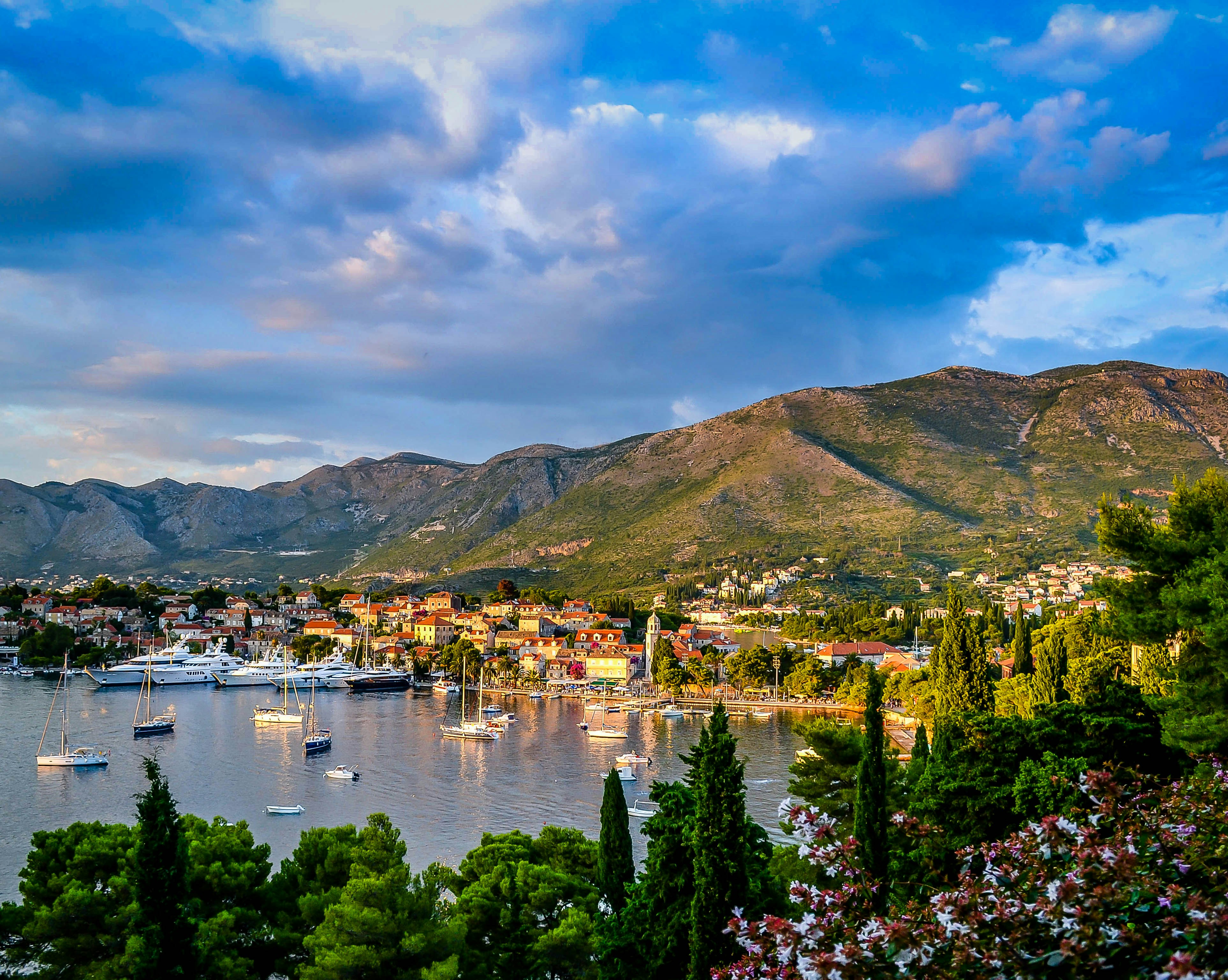 boats on body of water surrounded by trees and houses near mountain under blue and white sky at daytime