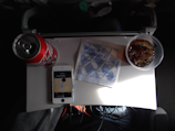 A small airplane tray table with a can of Coca-Cola, a plastic cup with ice and a dark beverage, an iPhone playing music, and a napkin with airline branding. The tray is part of an airplane seat, and the setting is dimly lit.