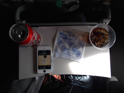 A small airplane tray table with a can of Coca-Cola, a plastic cup with ice and a dark beverage, an iPhone playing music, and a napkin with airline branding. The tray is part of an airplane seat, and the setting is dimly lit.
