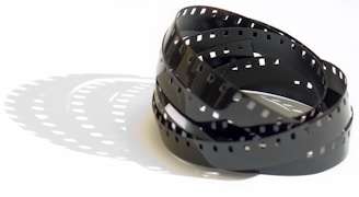 A coiled strip of photographic film rests on a white surface, casting distinct shadows that mirror its perforated edges and curving form. The film's glossy finish produces subtle reflections in the light.