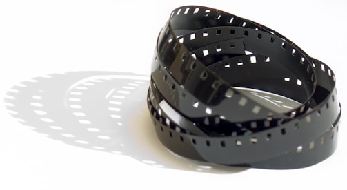 A coiled strip of photographic film rests on a white surface, casting distinct shadows that mirror its perforated edges and curving form. The film's glossy finish produces subtle reflections in the light.