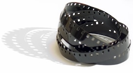 A coiled strip of photographic film rests on a white surface, casting distinct shadows that mirror its perforated edges and curving form. The film's glossy finish produces subtle reflections in the light.