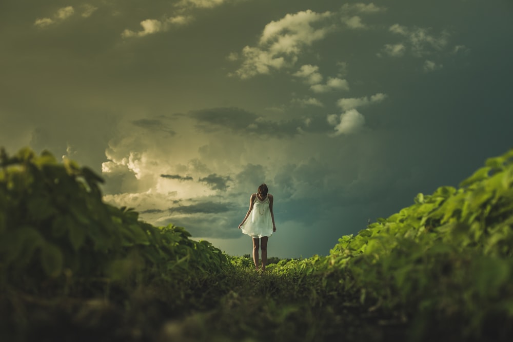 woman wearing white dress standing on hill with green grass under white cloudy sky