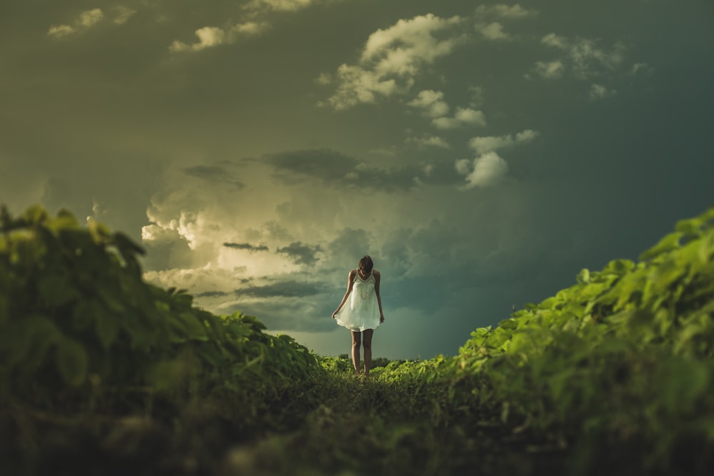 Frau im weißen Kleid steht auf einem Hügel mit grünem Gras unter weißem, bewölktem Himmel