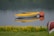 A yellow kayak is floating on a calm body of water, reflected smoothly on the surface. Next to the kayak, a large red buoy is visible. In the foreground, a strip of green grass is dotted with small yellow flowers, and the edge of another boat is partially visible.