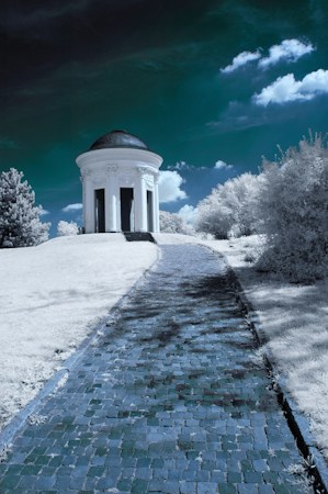 A cobblestone path leads to a small, round white pavilion with columns, set against a dramatic sky. The landscape, including grass and trees, appears desaturated or frosty, giving it a surreal and dreamlike quality.