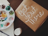 A flat lay featuring a paint palette with various colors, a brush, a paper or cardboard with the hand-painted phrase 'Get Out There' in white script, and an open jar of paint. The items are arranged on a dark surface, creating an artistic and inspiring setting.