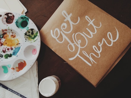 A flat lay featuring a paint palette with various colors, a brush, a paper or cardboard with the hand-painted phrase 'Get Out There' in white script, and an open jar of paint. The items are arranged on a dark surface, creating an artistic and inspiring setting.