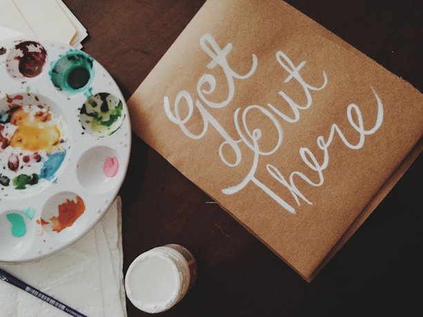 A flat lay featuring a paint palette with various colors, a brush, a paper or cardboard with the hand-painted phrase 'Get Out There' in white script, and an open jar of paint. The items are arranged on a dark surface, creating an artistic and inspiring setting.