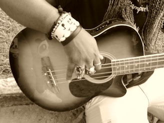A person is holding and playing a guitar, with a focus on their hand and the instrument's strings. The image is in sepia tone, and the person is wearing rings and bracelets. The guitar reflects part of a face sticker or design near the body.