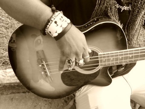 A person is holding and playing a guitar, with a focus on their hand and the instrument's strings. The image is in sepia tone, and the person is wearing rings and bracelets. The guitar reflects part of a face sticker or design near the body.