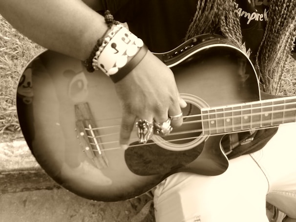 A person is holding and playing a guitar, with a focus on their hand and the instrument's strings. The image is in sepia tone, and the person is wearing rings and bracelets. The guitar reflects part of a face sticker or design near the body.