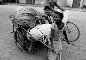 A young person is pulling a cart loaded with large bundles tied securely, walking on an urban street. In the background, a cyclist is riding past, creating a sense of bustling activity.