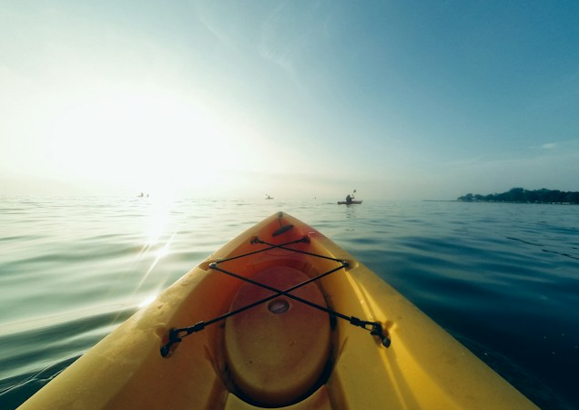 Bow of a yellow kayak on calm open water with the sun low and bright over the horizon and other paddlers in the distance