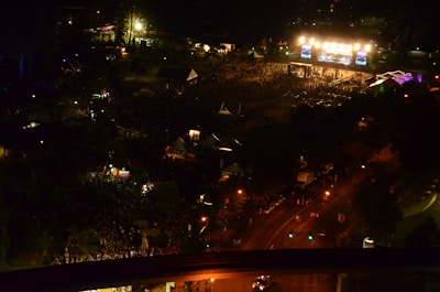A large outdoor nighttime event with numerous people gathered around a brightly lit stage area. The scene is bustling with activity, and surrounding tents and structures are visible as well as the illuminated roads leading towards the venue. Colorful lights and a festive atmosphere are prominent.