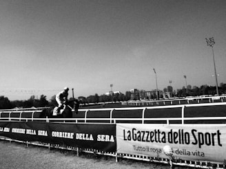 A horse and jockey race along a track, captured in black and white. The scene is framed by advertising banners on the side, including names of newspapers. A cityscape and trees are visible in the background under a clear sky.