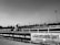 A horse and jockey race along a track, captured in black and white. The scene is framed by advertising banners on the side, including names of newspapers. A cityscape and trees are visible in the background under a clear sky.