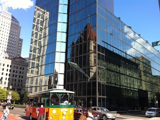 A modern glass building reflects various architectural structures, including a historic church with a pointed roof. An orange and green trolley bus travels along a busy city street, alongside pedestrians and cars. The street sign reads St. James Ave.