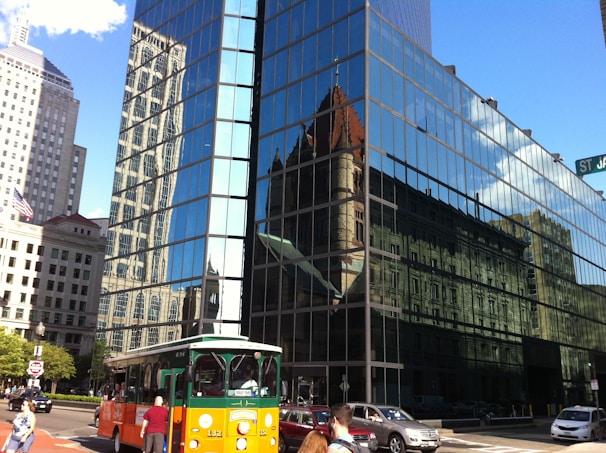 A modern glass building reflects various architectural structures, including a historic church with a pointed roof. An orange and green trolley bus travels along a busy city street, alongside pedestrians and cars. The street sign reads St. James Ave.