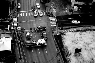 A black-and-white aerial view of a city intersection with multiple vehicles including police cars and a fire truck. The intersection appears to be cordoned off with police barricades, and several officers and personnel are visible. Traffic is halted, and the scene looks like an emergency or accident response.