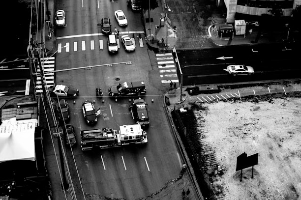 A black-and-white aerial view of a city intersection with multiple vehicles including police cars and a fire truck. The intersection appears to be cordoned off with police barricades, and several officers and personnel are visible. Traffic is halted, and the scene looks like an emergency or accident response.