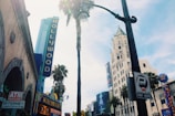 A vibrant urban scene with iconic Hollywood signage against a backdrop of tall buildings and palm trees. The atmosphere is bustling with various signs, including one for an ATM machine and internet access. A large sign for a wax museum is visible, as well as a parking meter sign. The sky is clear, suggesting a sunny day.