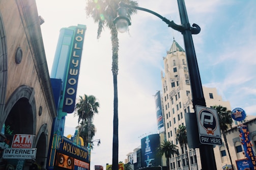 A vibrant urban scene with iconic Hollywood signage against a backdrop of tall buildings and palm trees. The atmosphere is bustling with various signs, including one for an ATM machine and internet access. A large sign for a wax museum is visible, as well as a parking meter sign. The sky is clear, suggesting a sunny day.