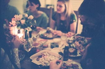 A cozy, dimly lit gathering around a dinner table where several people are engaged in conversation and enjoying a meal. The table is adorned with candles, flowers, and a variety of dishes, creating a warm and inviting atmosphere.