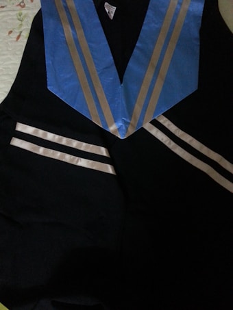 A black graduation gown with a blue stole featuring silver stripes laying on a surface.