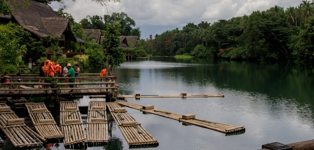 A tranquil riverside scene with thatched-roof huts nestled among lush greenery. Wooden rafts float on the calm water, and several people are gathered on a wooden dock with life vests hanging nearby. The trees on the riverbank create a natural, serene atmosphere.