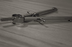 A metallic compass used for drawing circles or arcs lies on a wooden surface. The image focuses on the detailed parts of the compass, such as the hinge, adjusting screws, and pencil holder.