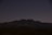A mountainous silhouette is visible against a dark, starry night sky. The landscape below appears rugged, with sparse vegetation typical of a desert environment.