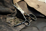 A pair of large, metal scissors placed on a rough, dark brown leather surface. Surrounding the scissors are several metal grommets and tools intended for leatherwork or crafting. The leather in the background has a worn texture, with some pieces appearing frayed or torn.