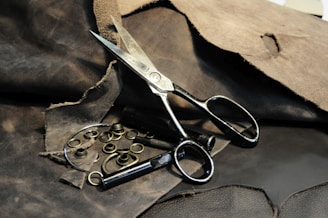 A pair of large, metal scissors placed on a rough, dark brown leather surface. Surrounding the scissors are several metal grommets and tools intended for leatherwork or crafting. The leather in the background has a worn texture, with some pieces appearing frayed or torn.